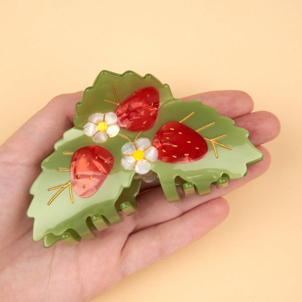 Pinza de pelo fresas Coucou Suzette sostenida en la palma de una mano sobre fondo melocotón