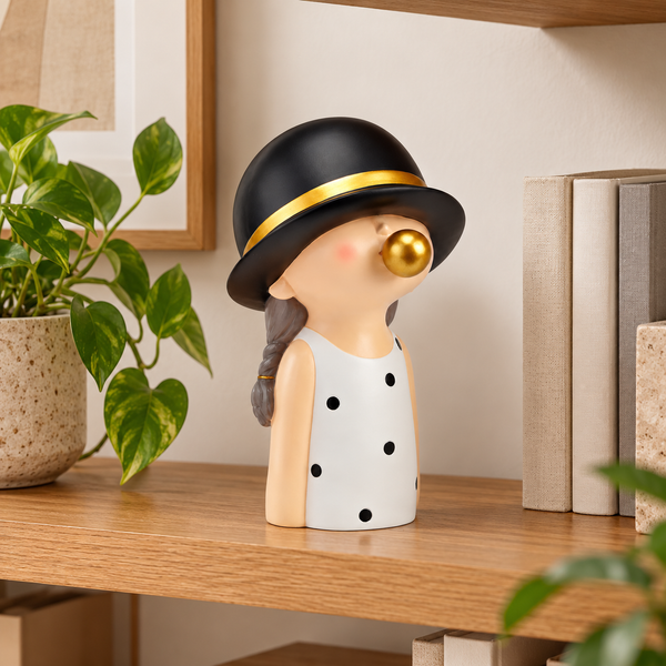 Figura decorativa de niña con chicle dorado colocada en estantería junto a libros y plantas.