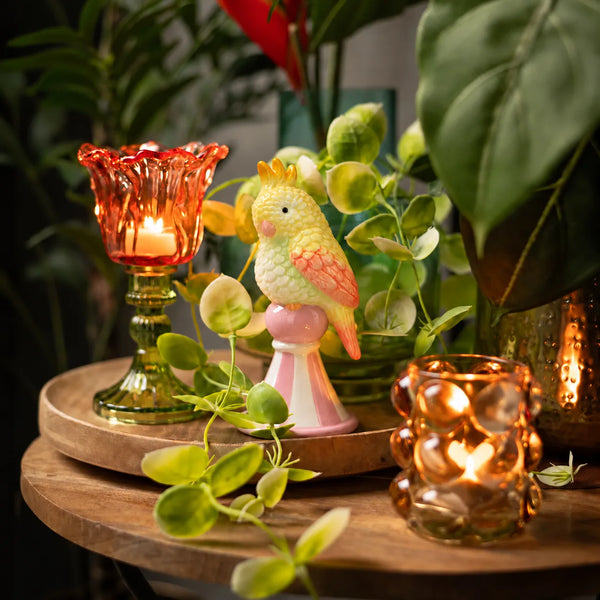 Figurita Loro Amarillo decorativa entre plantas y portavelas de cristal sobre bandeja de madera.