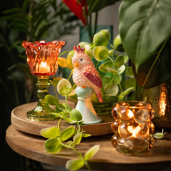 Figurita Loro Rosa decorativa entre plantas y portavelas sobre una bandeja de madera.