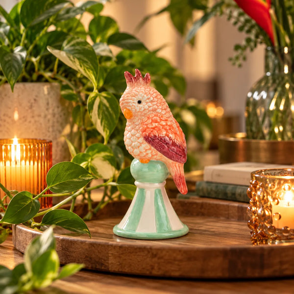 Figurita Loro Rosa de cerámica integrada en una composición decorativa con plantas y luz cálida.