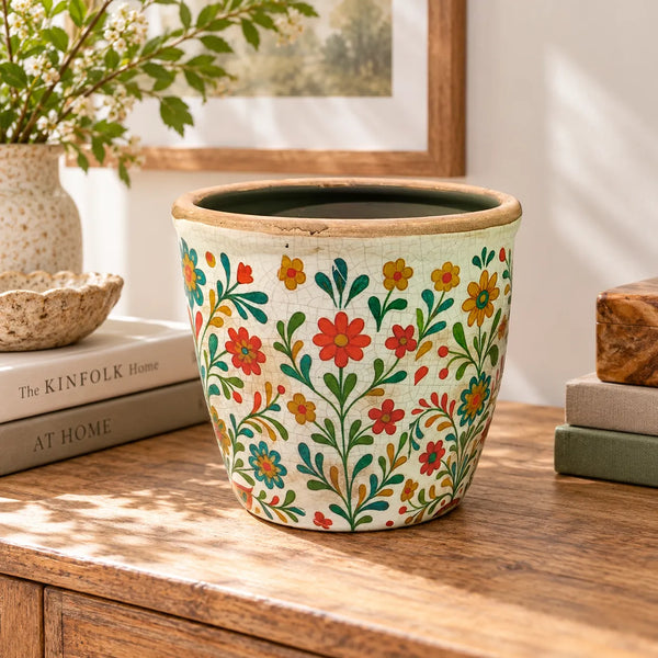 Macetero decorativo con estampado floral craquelado sobre cómoda de madera junto a libros y jarrones.