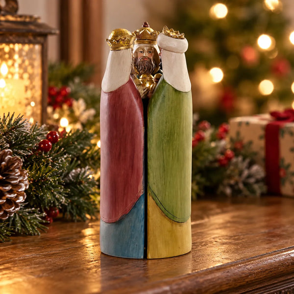 Vista posterior de nacimiento tríptico de los Reyes Magos cerrado, con mantos en tonos rojo, verde y amarillo y coronas doradas, sobre una mesa decorada para Navidad.